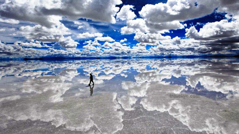 A stunning wide-angle shot of a person standing on the Uyuni Salt Flats in Bolivia, perfectly reflected in the shallow water, creating a surreal mirror effect