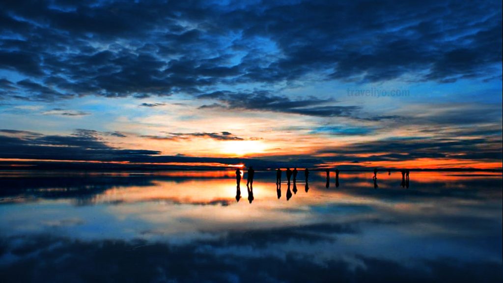 A beautiful sunset over the Uyuni Salt Flats with two figures silhouetted against the vibrant orange and blue sky, with the reflections on the water
