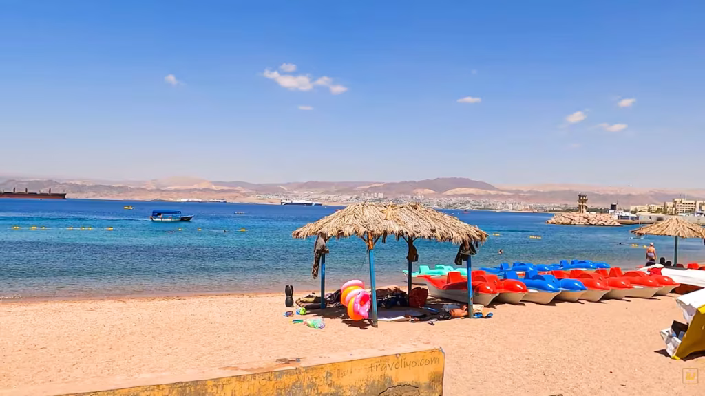 Straw umbrellas and colorful small boats resting on the sandy public beach in Aqaba, Jordan.