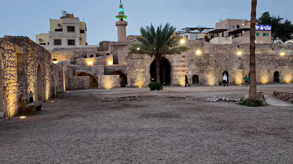 The illuminated stone courtyard and historic walls of the Aqaba Mamluk Fort at dusk with a mosque minaret in the background.