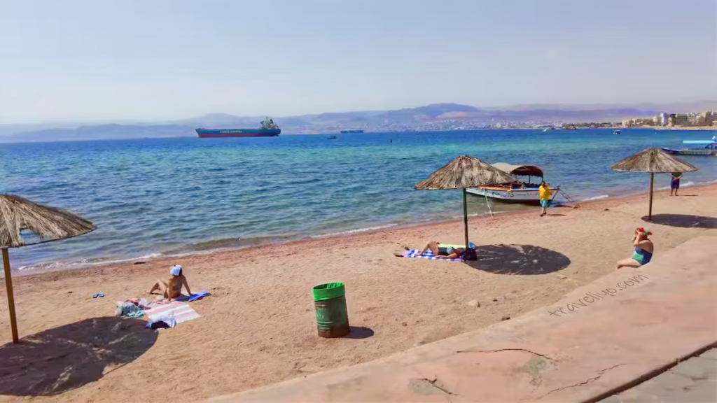 Tourists relaxing on the Aqaba beach looking out towards the blue waters of the Red Sea and distant ships.