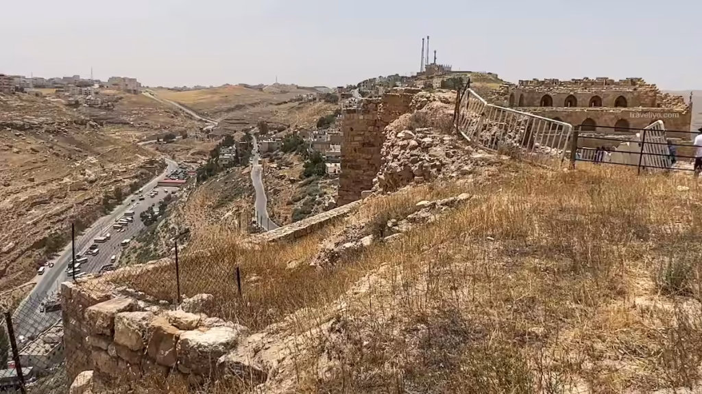 High outer defensive stone walls of Karak Castle overlooking a modern winding road in the valley below.