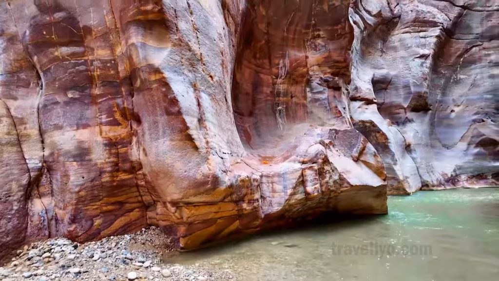 Text: Vibrant orange and pink rock formations reflecting in the water inside Wadi Mujib.