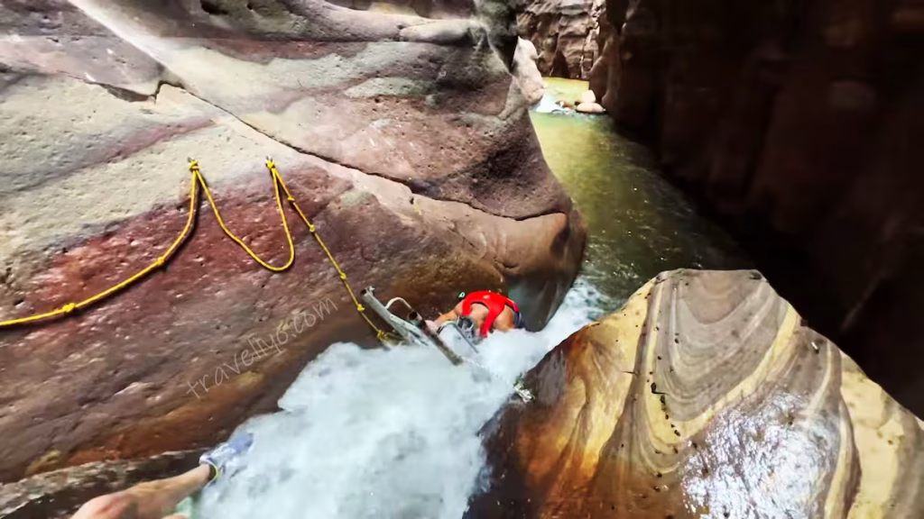 A hiker climbing up a metal ladder and using guide ropes against a rushing waterfall on the Wadi Mujib Siq Trail.
