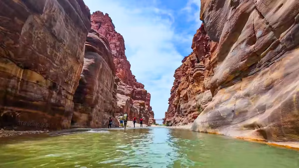A group of small hikers walking through the bottom of the immense vertical cliffs of Wadi Mujib.