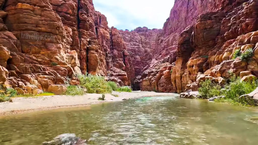 Towering red sandstone cliffs framing the rushing river inside a canyon in Wadi Mujib.