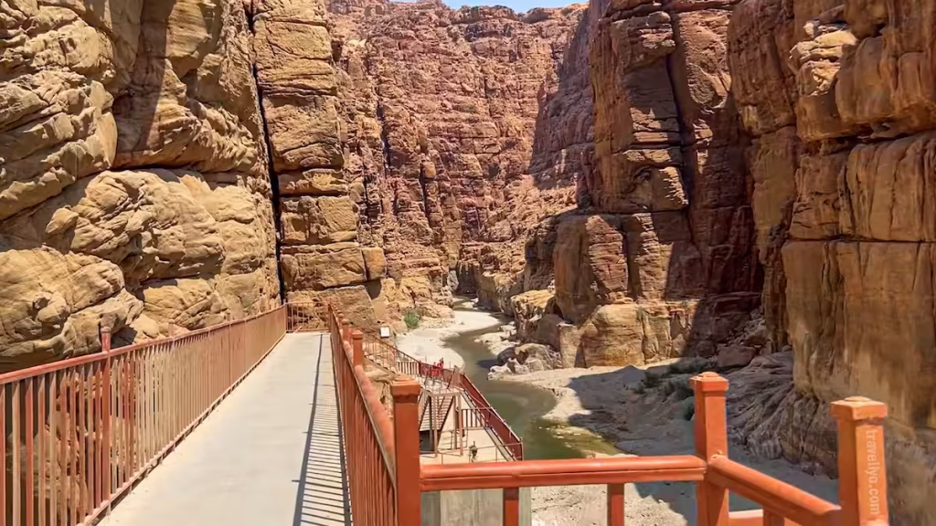A metal walkway structure leading into the massive sandstone cliffs of Wadi Mujib near the Dead Sea.